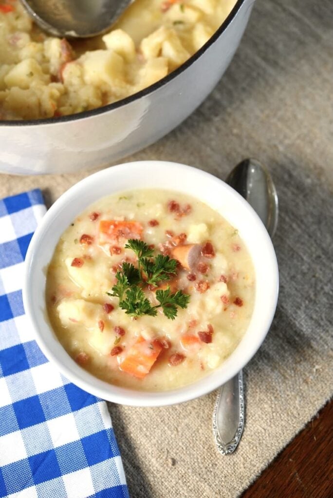 A bowl of potato soup topped with bacon bits and parsley, with a large pot of the same soup in the background.