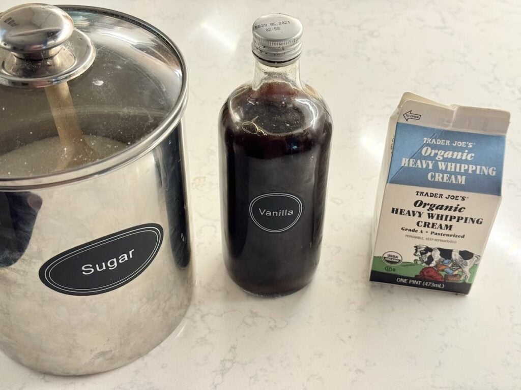 A metal sugar canister, a glass bottle of vanilla extract, and a carton of Trader Joes organic heavy whipping cream on a white countertop.