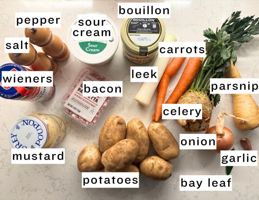 Ingredients for potato soup arranged on a countertop, labeled with salt, pepper, sour cream, bouillon, wieners, bacon, leek, carrots, celery root, parsnip, mustard, potatoes, onion, garlic, and bay leaf.