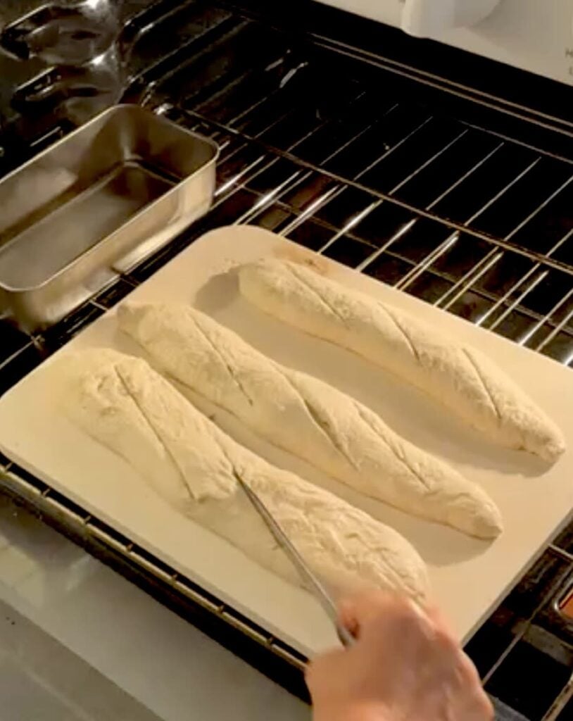 A hand uses a lame to score the tops of three raw sourdough baguette loaves on a baking stone inside an oven.