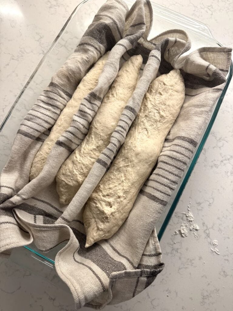 Three shaped sourdough-baguette loaves of raw dough resting in a glass dish, separated by folds in a striped cloth.