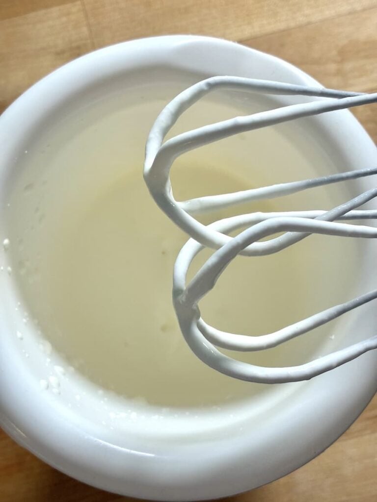 A wire whisk held over a white bowl filled with a thin white liquid.