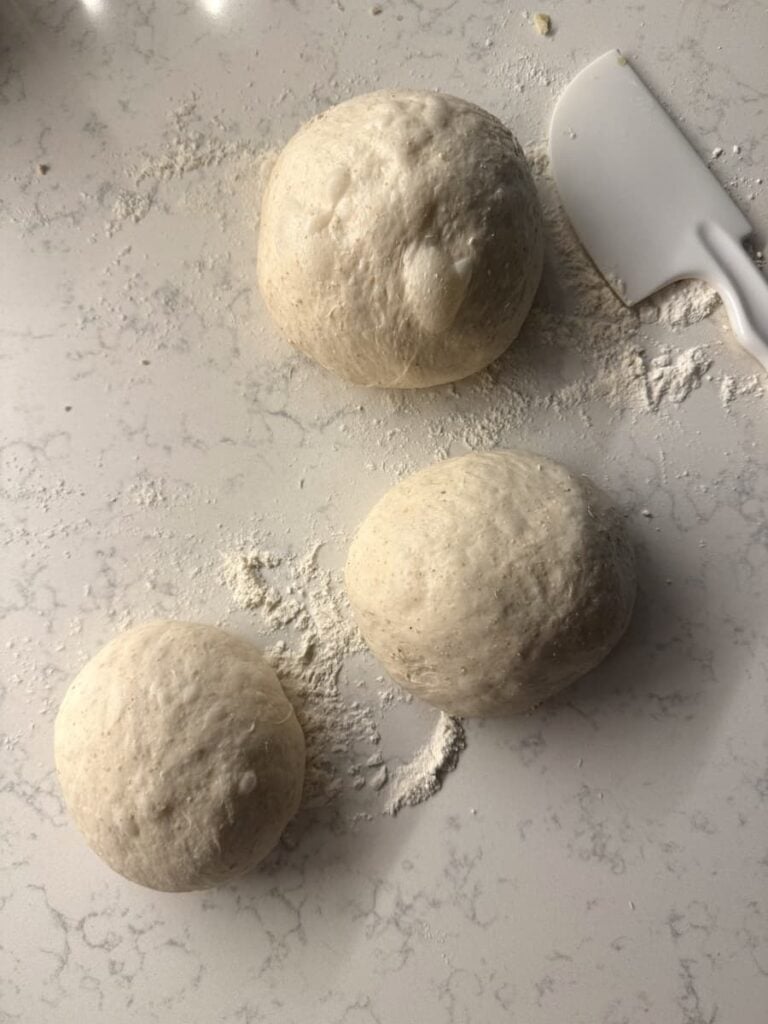 Three rounds of proofed sourdough-baguette dough on a floured countertop with a plastic bench scraper nearby.