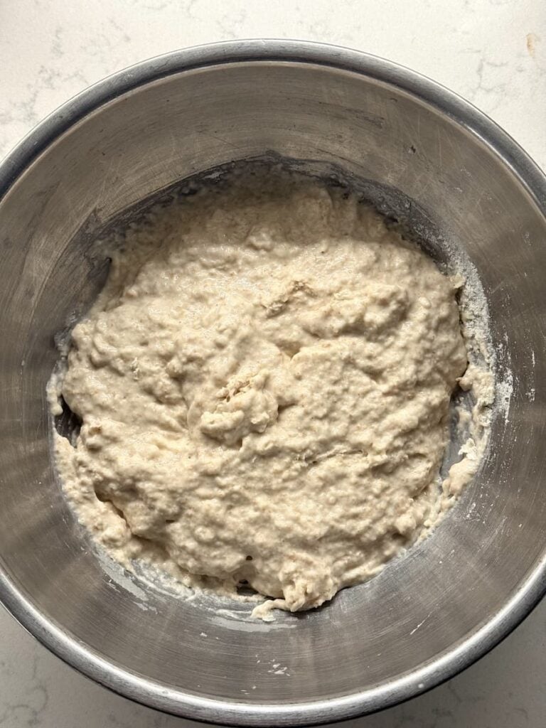 A metal mixing bowl containing a ball of shaggy, raw sourdough baguette dough.