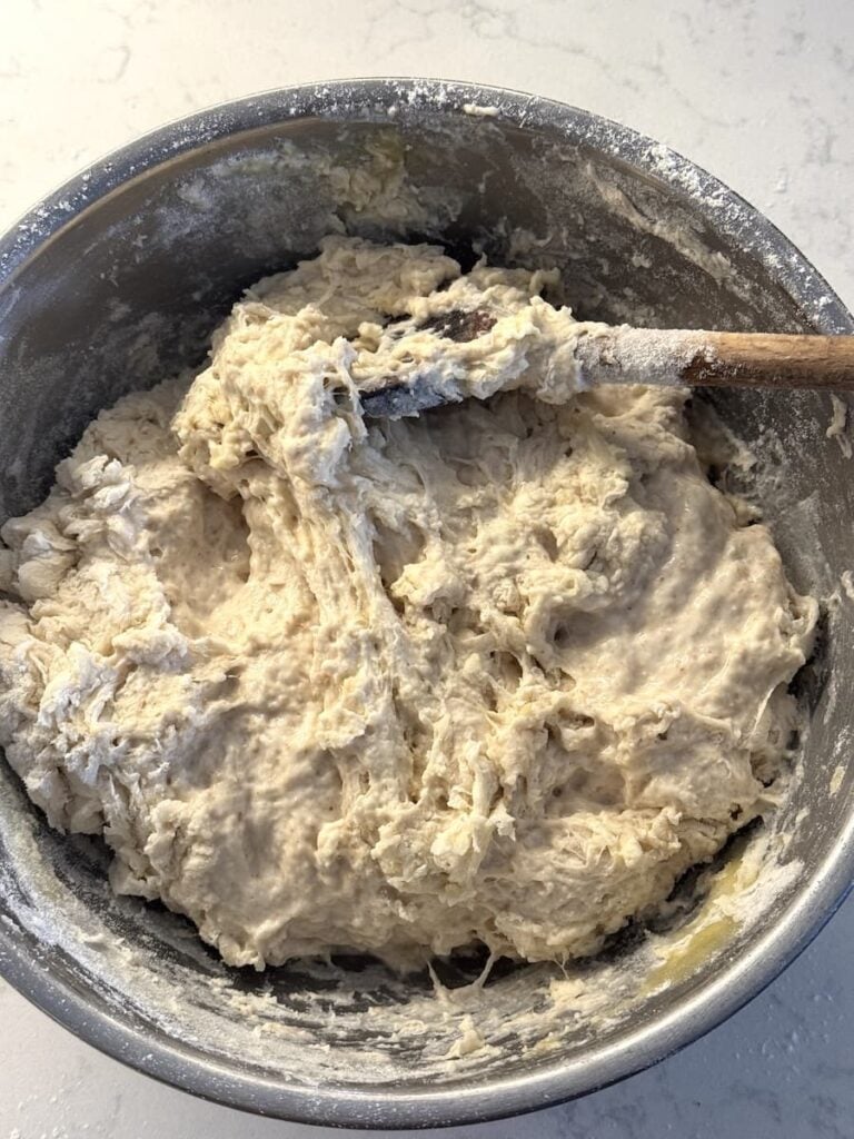 A wooden spoon stirs shaggy, unkneaded sourdough baguette dough in a metal mixing bowl.