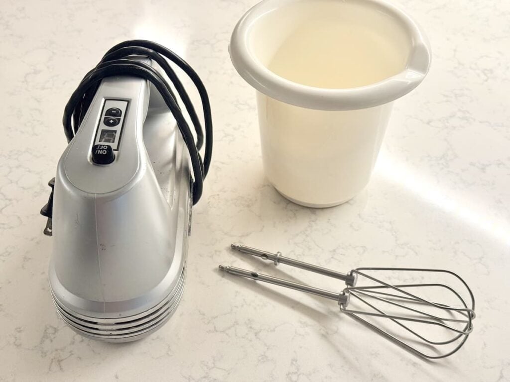 A silver electric hand mixer, a white plastic mixing bowl, and two wire beaters on a countertop.