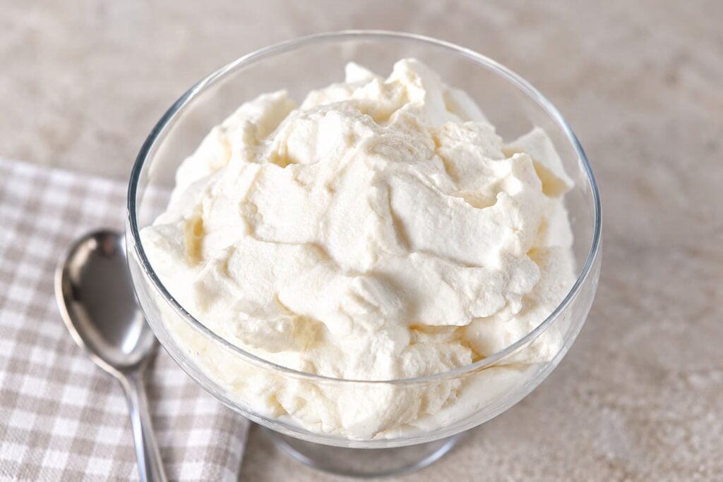 A glass bowl filled with whipped cream, next to a silver spoon on a checkered napkin.