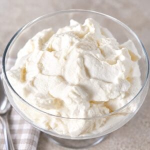 A glass bowl filled with whipped cream, next to a silver spoon on a checkered napkin.