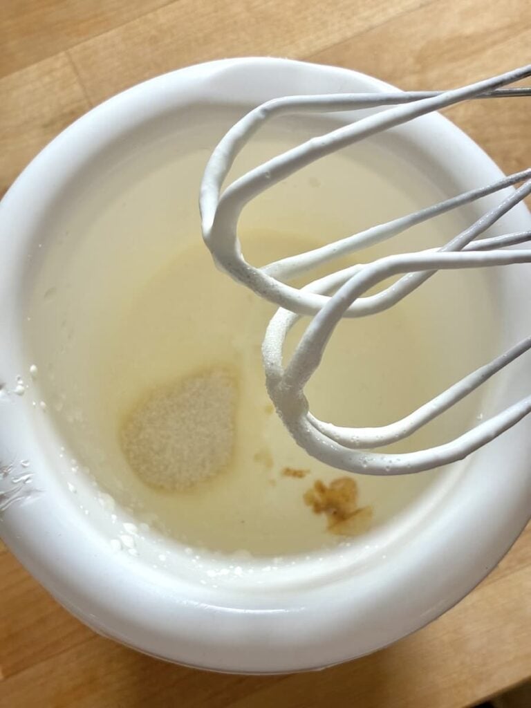 White mixing bowl containing liquid, sugar, and vanilla extract with a wire whisk held above it.